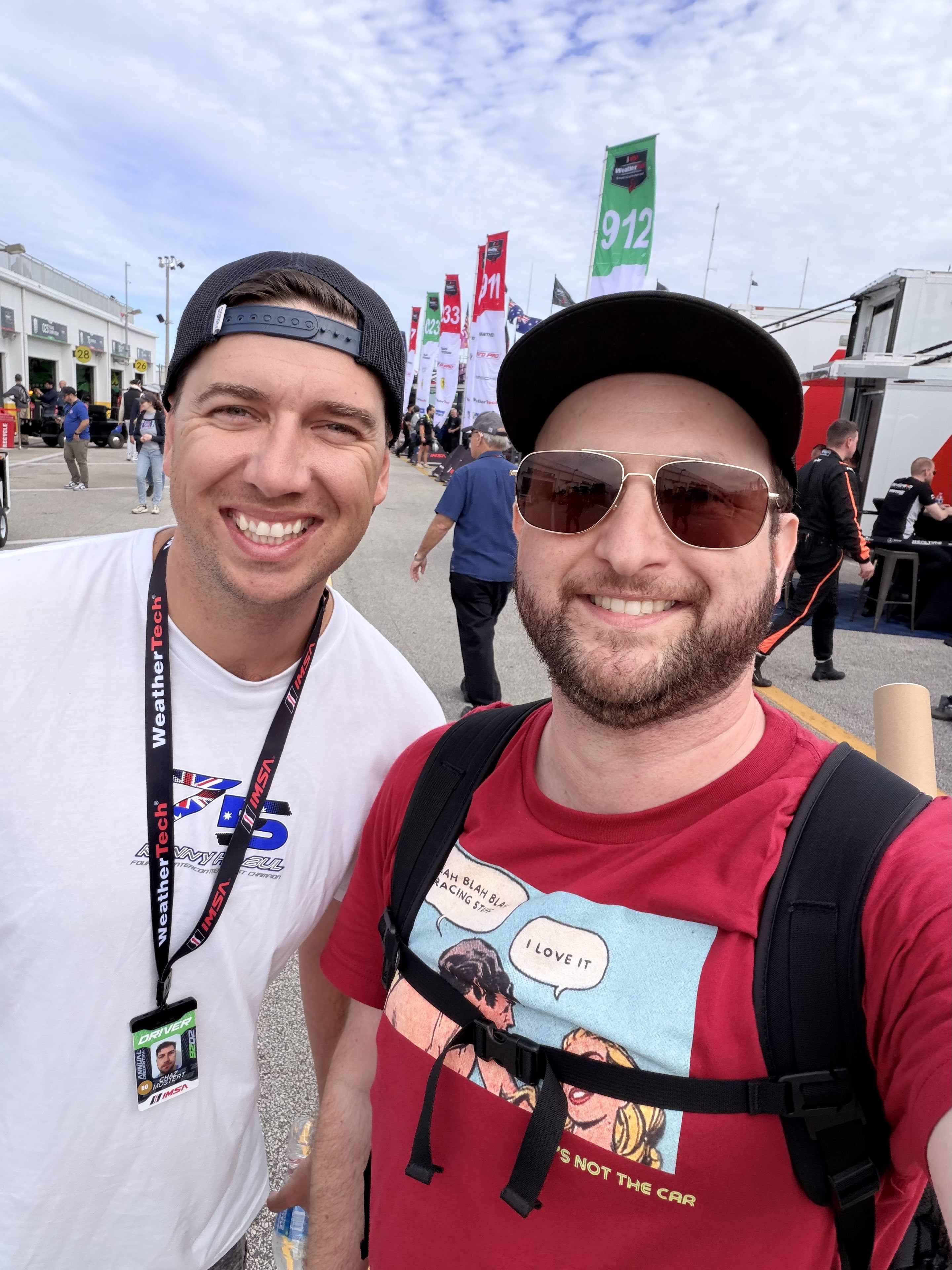 Two men smiling in the garage area of a motor race, one with his hat forwards, the other with his hat backwards and a credential around his neck